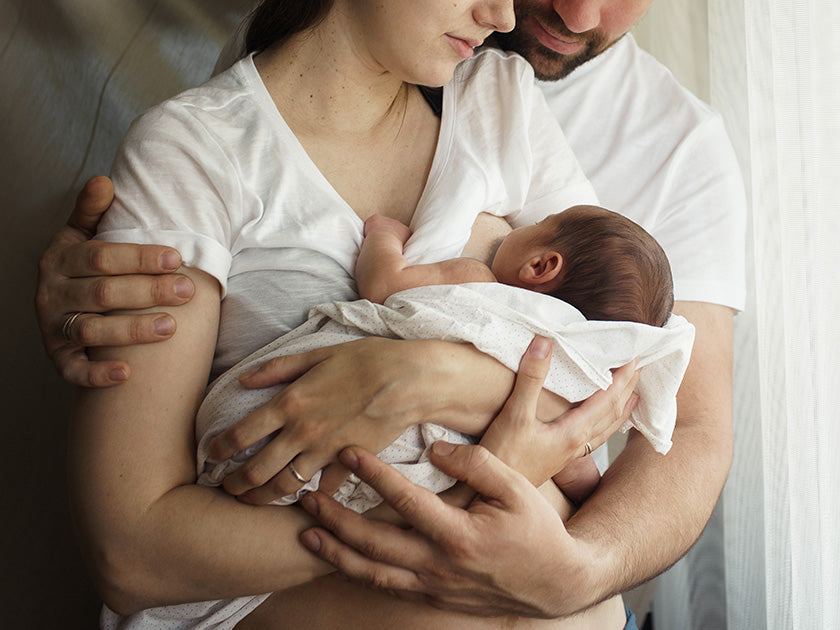 Un bébé emmailloté dans une couverture, tenu dans les bras de sa mère, avec le père qui les entoure, en arrière-plan une fenêtre lumineuse.