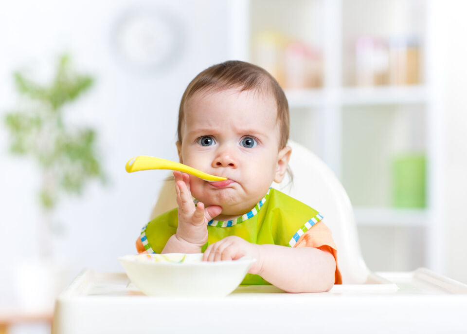 Un bébé assis dans une chaise haute, l'air perplexe, tient une cuillère jaune dans la bouche.