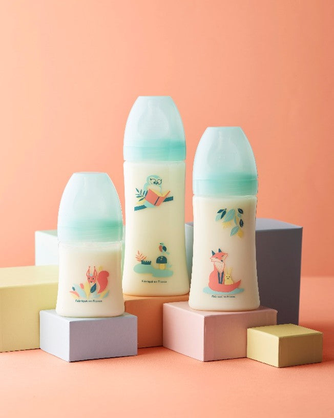 Trois biberons pour bébé aux bouchons bleu clair, décorés d’illustrations colorées d’animaux et de nature, posés sur des cubes pastel sur un fond orange