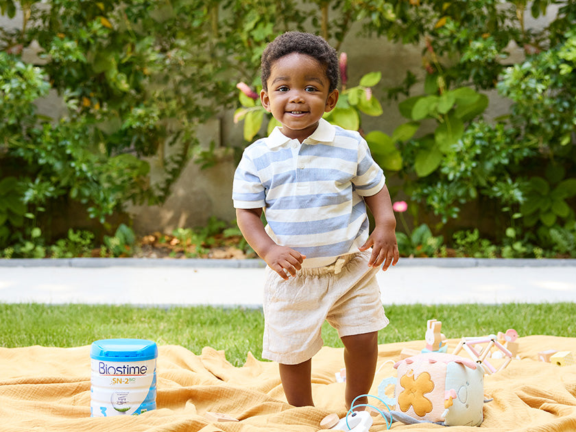 Un jeune garçon souriant, debout sur une couverture jaune dans un jardin. À côté de lui, une boîte de lait infantile 