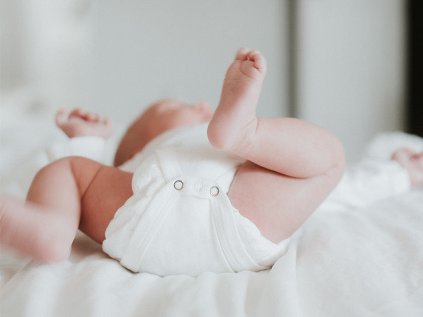 Un bébé allongé sur le dos, portant une couche blanche, avec ses jambes levées en l’air dans un environnement doux et lumineux.