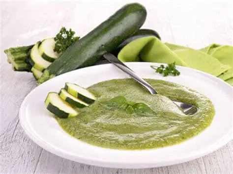 Assiette blanche contenant une purée verte de courgette, garnie de tranches de courgette et décorée de feuilles de basilic, avec une cuillère posée dans l'assiette