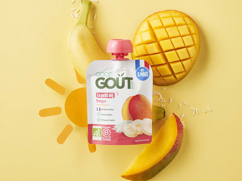 Gourde de compote Good Goût à la mangue, posée sur un fond jaune avec des fruits frais : une banane, une mangue coupée en morceaux, le tout décoré d’un soleil dessiné.