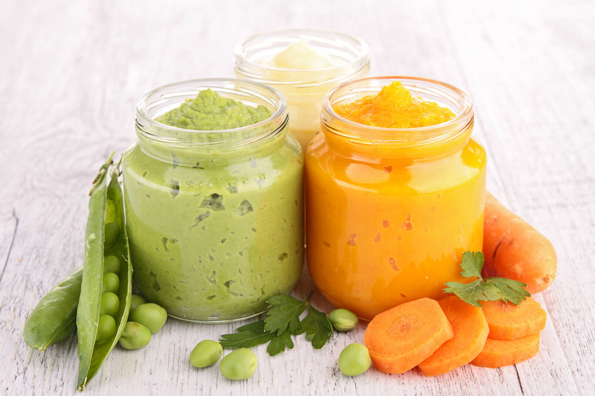 Des pots de purées colorées, à base de pois, carottes et autres légumes, disposés sur une surface en bois avec des tranches de carottes et des gousses de pois en arrière-plan. 