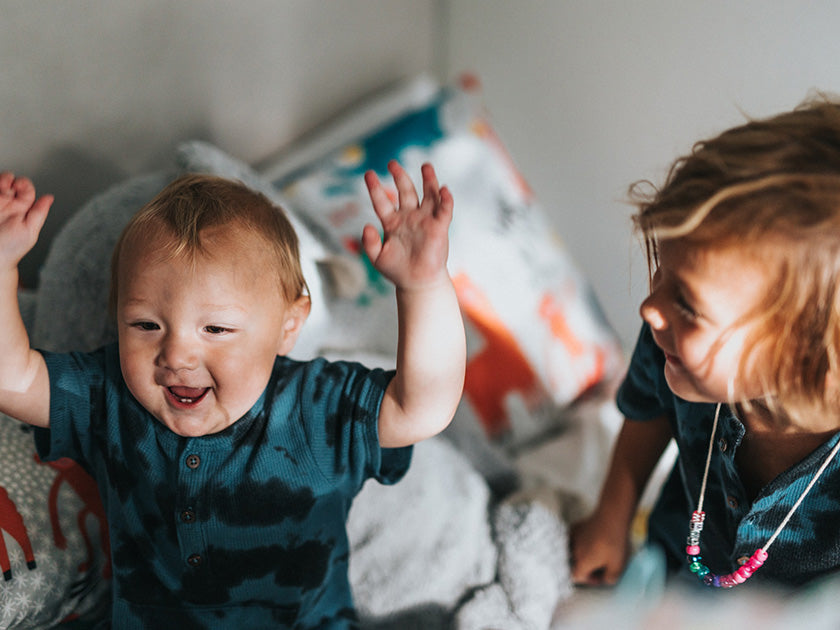 Deux jeunes enfants jouent ensemble sur un lit, l’un d’eux lève joyeusement les bras en l’air.