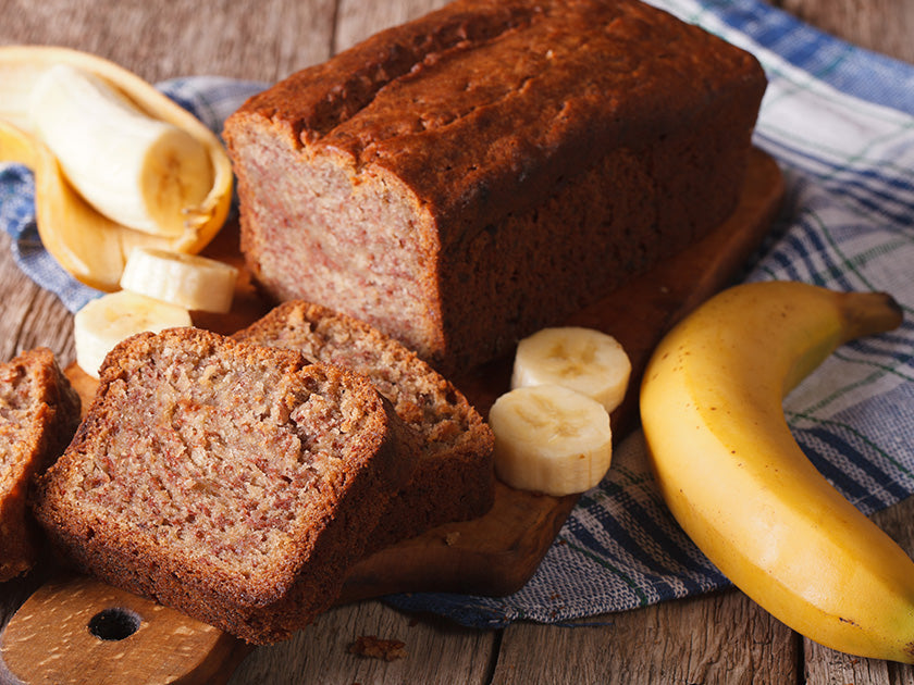 Un cake à la banane tranché posé sur une planche en bois, entouré de morceaux de banane pelée et d’une banane entière, sur un tissu à carreaux.