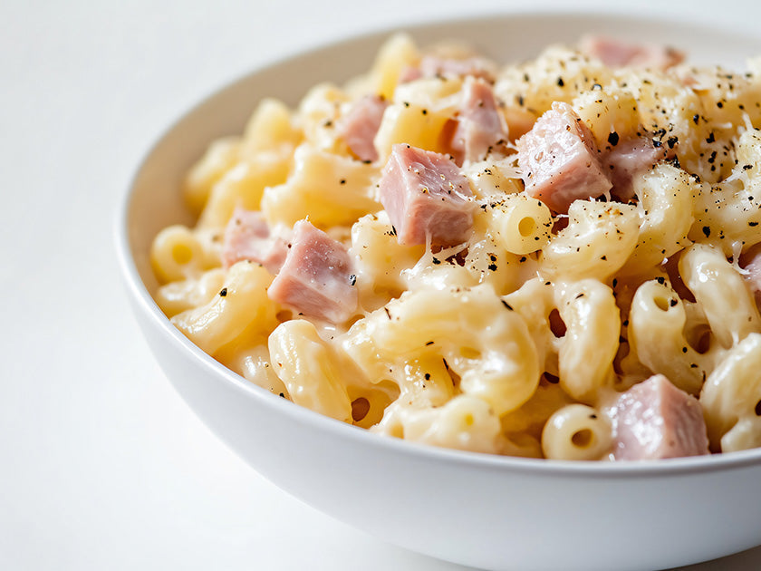 Bol blanc rempli de macaroni au fromage crémeux avec des morceaux de jambon et une pincée de poivre noir fraîchement moulu.