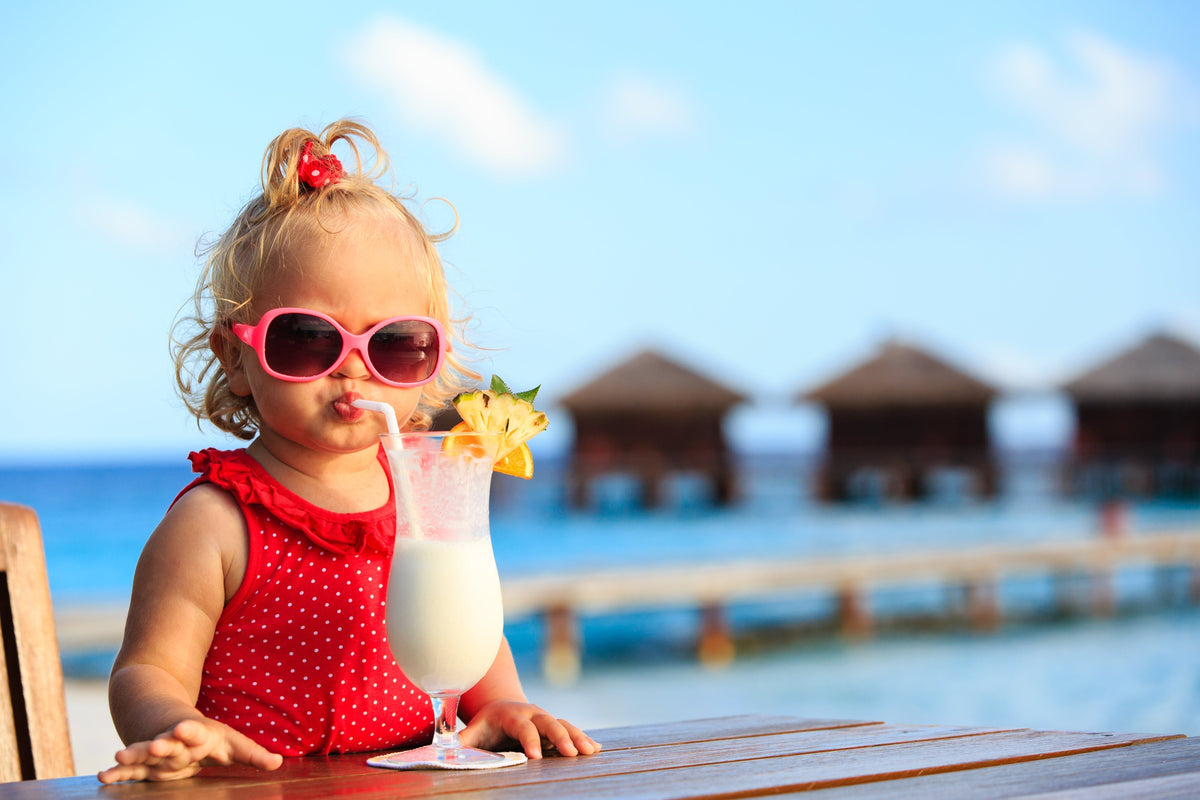 Petite fille en lunettes roses sirotant un cocktail en bord de mer, avec des bungalows sur pilotis en arrière-plan.