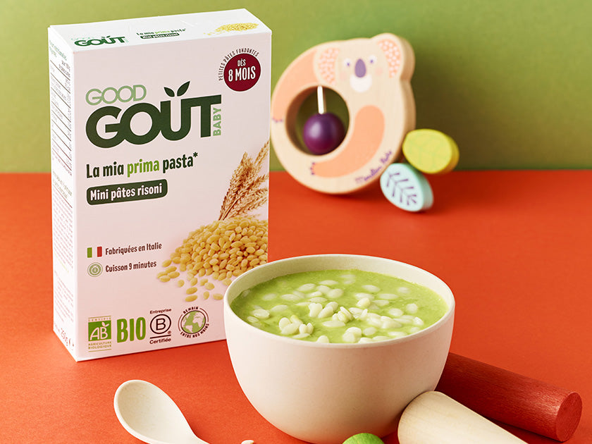 Boîte de pâtes bio Good Goût pour bébé à partir de 9 mois, accompagnée d’un bol de soupe verte avec des petites pâtes en forme d’étoiles, sur fond orange, avec un jouet pour bébé à côté.